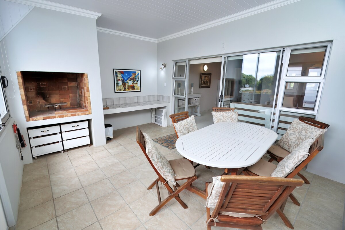 An outdoor dining area is presented, featuring a round white table surrounded by six wooden chairs with light fabric cushions. A built-in barbecue is located adjacent to the dining space, with a counter and cabinets visible against a neutral wall.