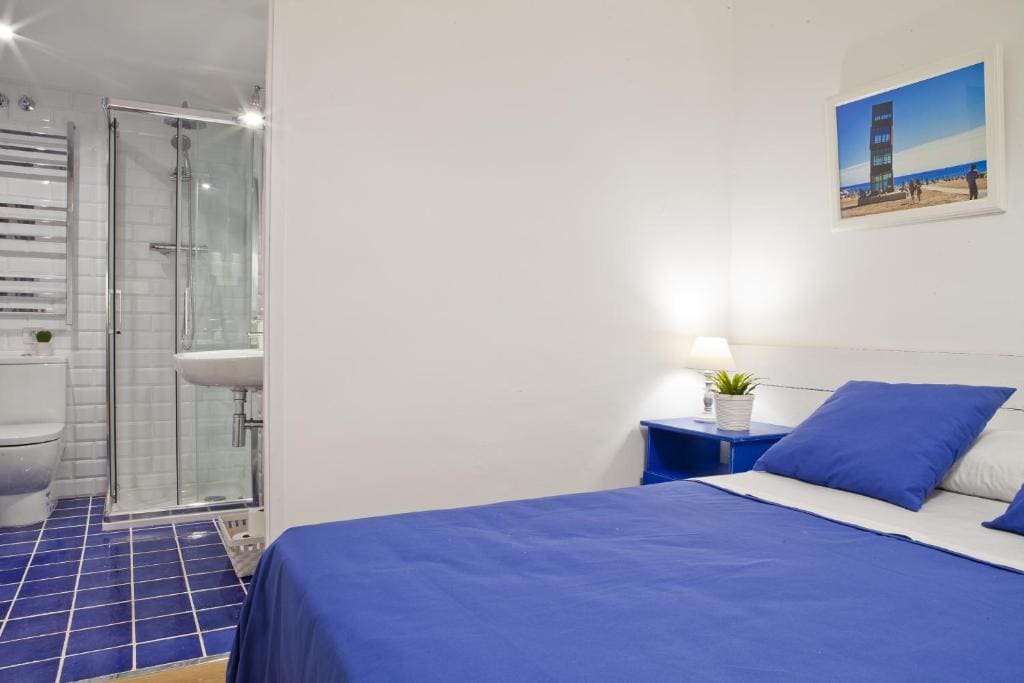 The room features a double bed adorned with blue bedding, complemented by a bedside lamp and a small potted plant. A bright, modern bathroom is visible, showcasing a glass-enclosed shower and sleek fixtures. Light-colored walls enhance the airy feel of the space.