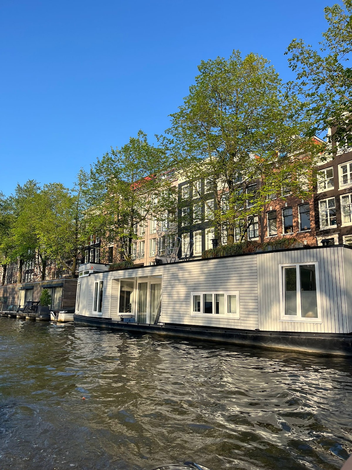 The image captures a scenic view of a residential area along a canal, featuring a modern houseboat with large windows. Lush trees frame the exterior, while the calm water reflects the clear blue sky and surrounding architecture.