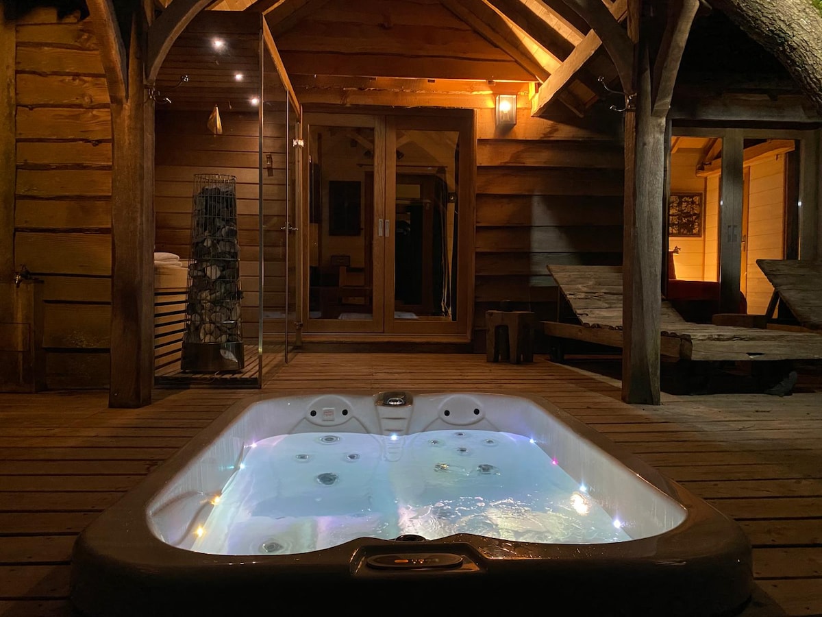 A private hot tub is located on a wooden deck, illuminated by soft lights. The surrounding wooden structure complements the relaxing ambiance, with glass doors leading into the cabin. Outdoor seating is positioned nearby, adding to the serene atmosphere.