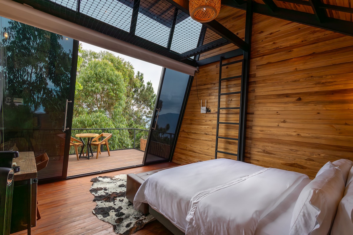 A spacious bedroom features large glass doors that open to a private deck, enhancing natural light. The wooden walls create a warm atmosphere, while a comfortable bed is complemented by a textured area rug and contemporary light fixtures.