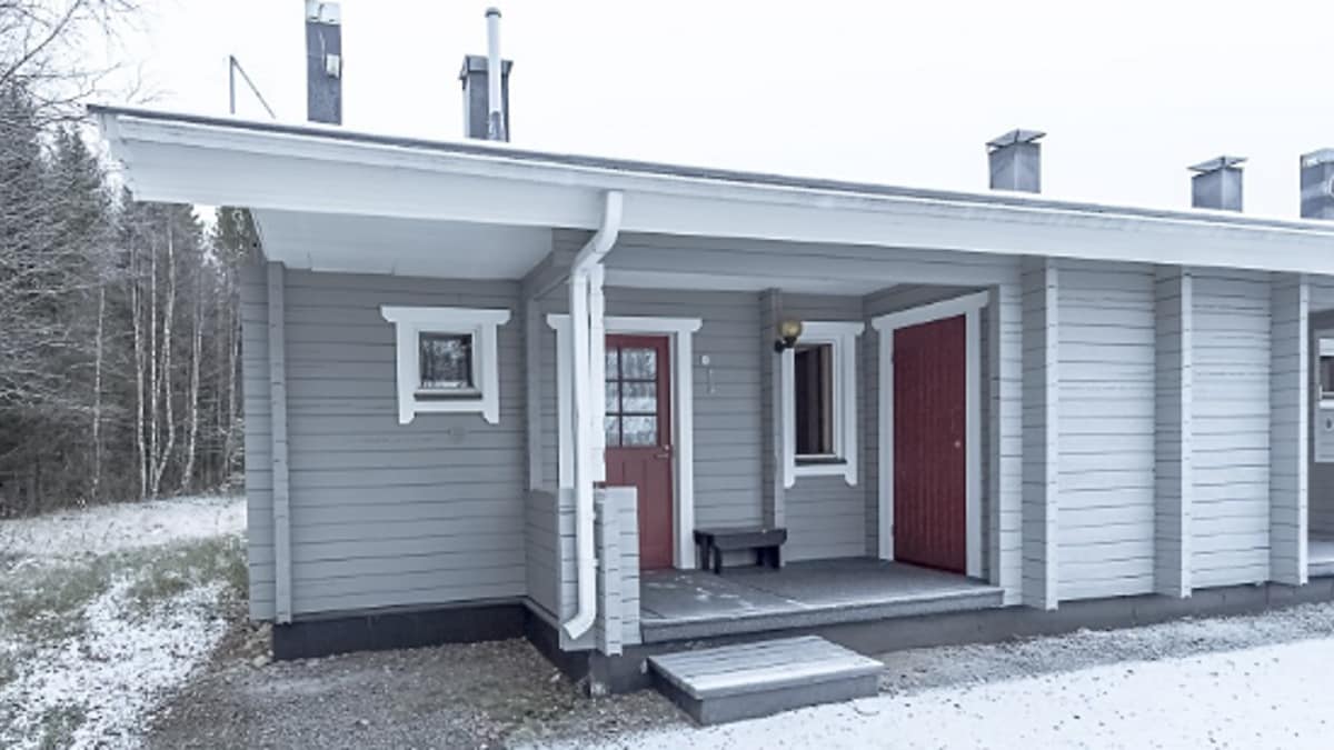 The exterior of a single-story corner apartment is shown, featuring a light gray wooden facade. A red front door and a matching side door are visible, along with a small covered porch and surrounding snow-covered ground. Several chimneys are seen above the roofline.