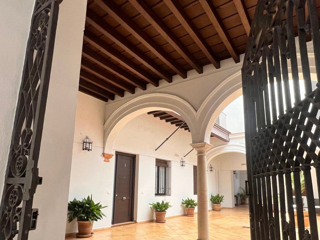 An inviting entryway is framed by grand arches and a natural wood ceiling. Potted plants add greenery to the space, while two lanterns provide subtle illumination. A door, marked with the number '2', leads into the apartment, enhancing the welcoming ambiance of the corridor.