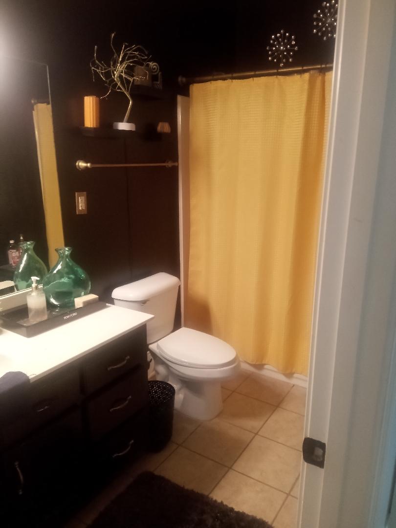 A modern bathroom is presented with dark walls and warm-toned tiles. A white toilet sits adjacent to a dark wooden vanity holding decorative items. A large mirror reflects the space, while a yellow shower curtain adds a pop of color, enhancing the overall ambiance.