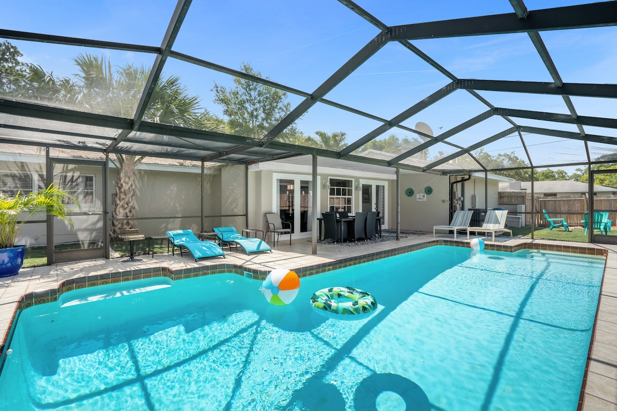 A private screened pool area features an inviting blue pool with lounge chairs and a dining table visible around the edge. Bright beach accessories add a playful touch, while a spacious yard with greenery is present in the background.