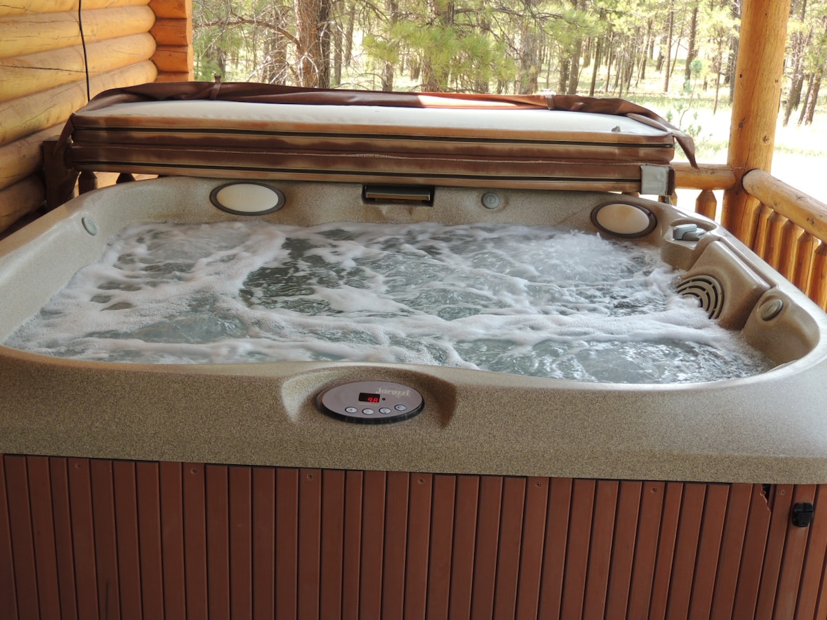 A hot tub is situated on a wooden deck, featuring bubbling water and adjustable jets. The tub is equipped with control buttons for temperature and settings. Surrounding trees provide a natural backdrop, enhancing the tranquil atmosphere of the space.