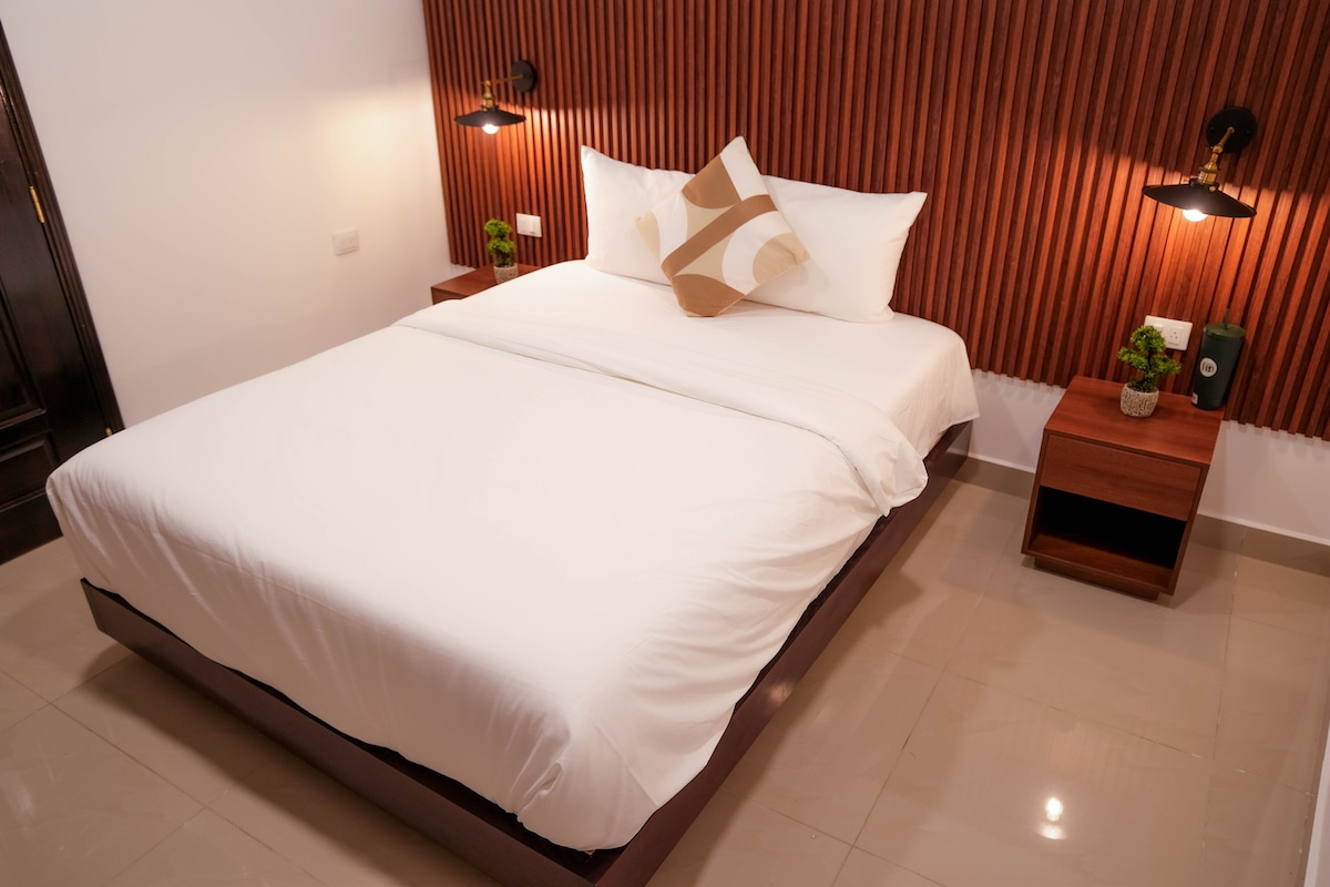 A spacious bed is centered against a textured wooden accent wall. Soft lighting is provided by bedside lamps on either side. Small potted plants and a minimalist nightstand complement the cozy ambiance, while smooth tiles extend across the floor.