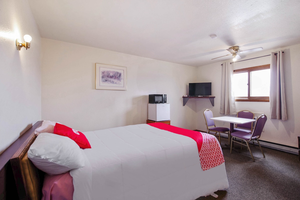 A clean, well-lit room is presented with a spacious bed covered in white linens. A small table with two chairs is visible, along with a television and a microwave. Natural light filters in through a window dressed with light curtains.