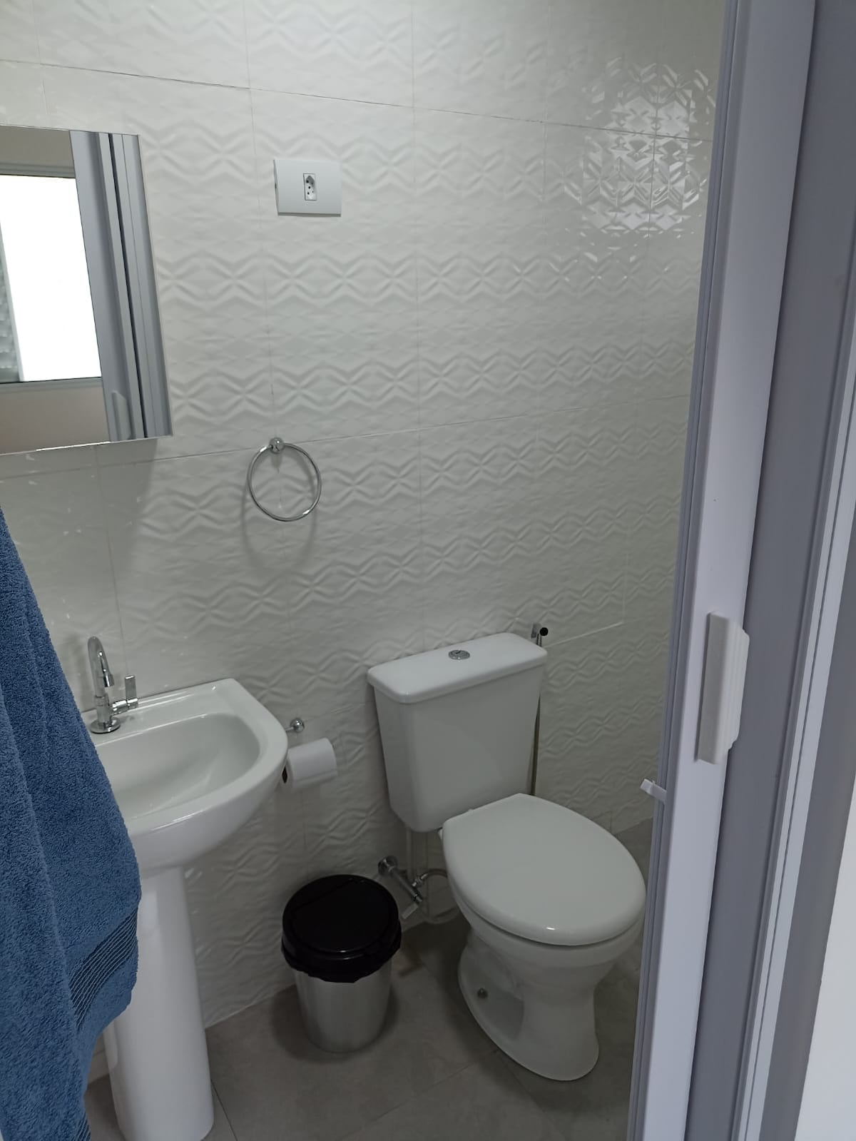 A compact bathroom is showcased, featuring a white sink with a mirror above, a toilet, and a small trash can. A blue towel is neatly hung on a wall-mounted ring, complementing the clean, tiled surfaces.