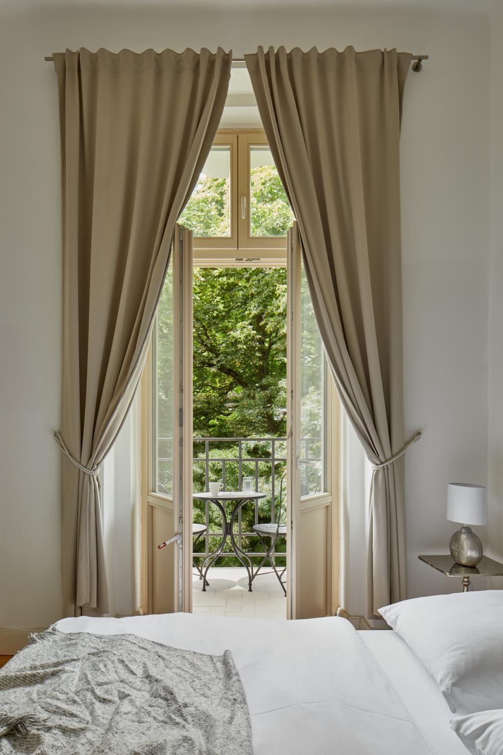 Large windows framed by light-colored curtains lead to a private balcony. The view showcases lush greenery, enhancing the room's tranquil feel. A cozy bed is visible, with a soft blanket casually draped over it, contributing to the inviting ambiance.