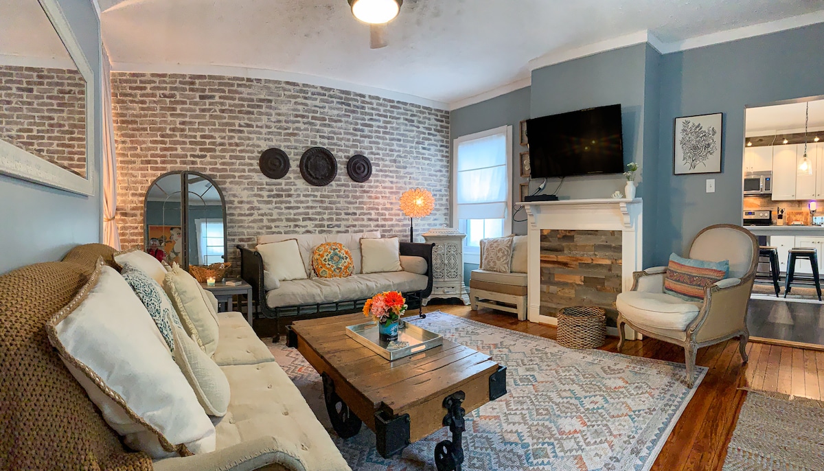 The living area features a comfortable seating arrangement with a neutral-colored sofa and an accent chair. A textured area rug sits beneath a rustic coffee table. Light from nearby windows illuminates the room, complemented by a modern fireplace and a wall-mounted television.