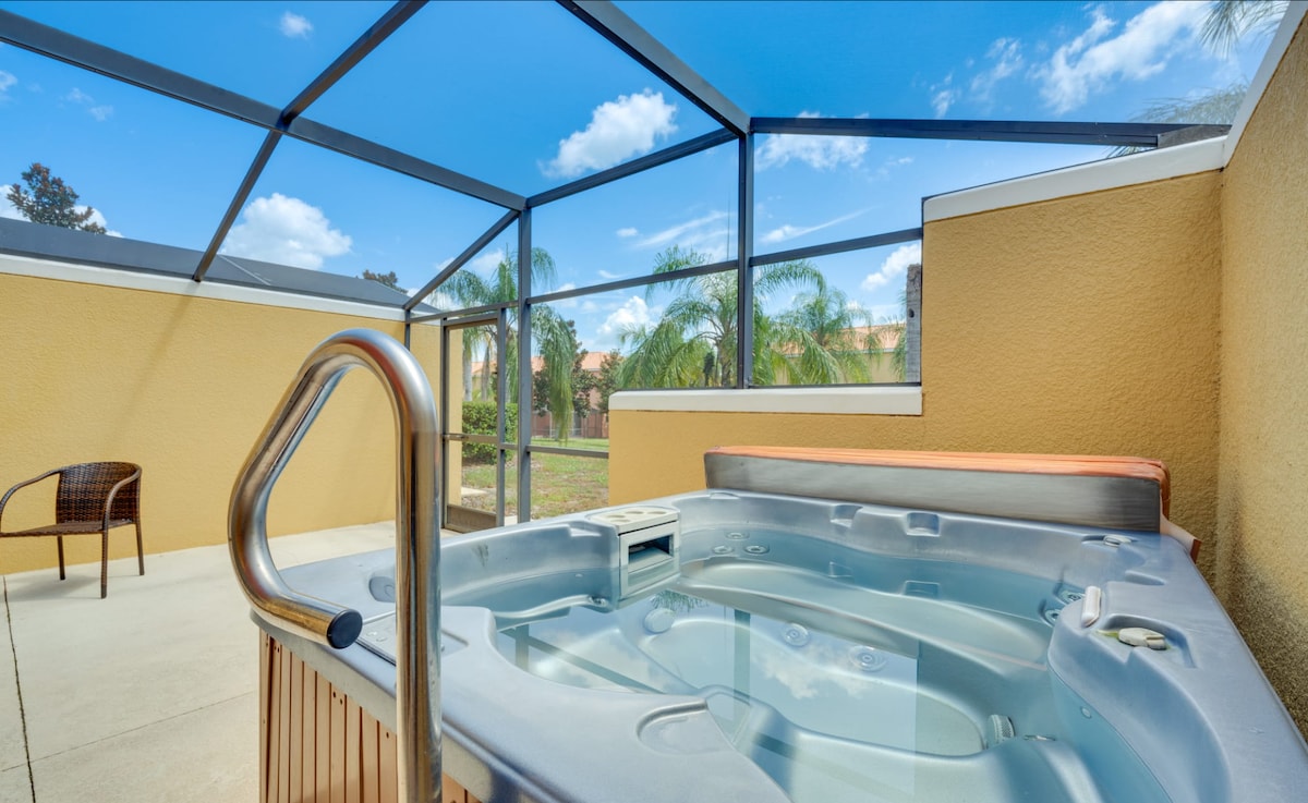 A private screened patio features a four-person hot tub, surrounded by a concrete surface. The vibrant blue sky is visible through the screening, while palm trees can be seen in the background, enhancing the outdoor experience.