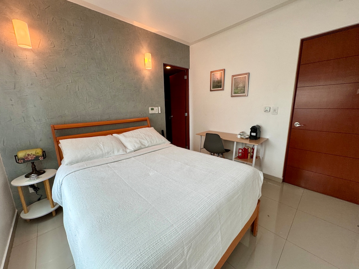 A comfortable room is showcased, featuring a spacious bed with white linens. A modern pendant light and wall art add subtle decoration. A small nightstand and a chair are visible, along with a kitchenette area equipped with a coffee maker and essentials.