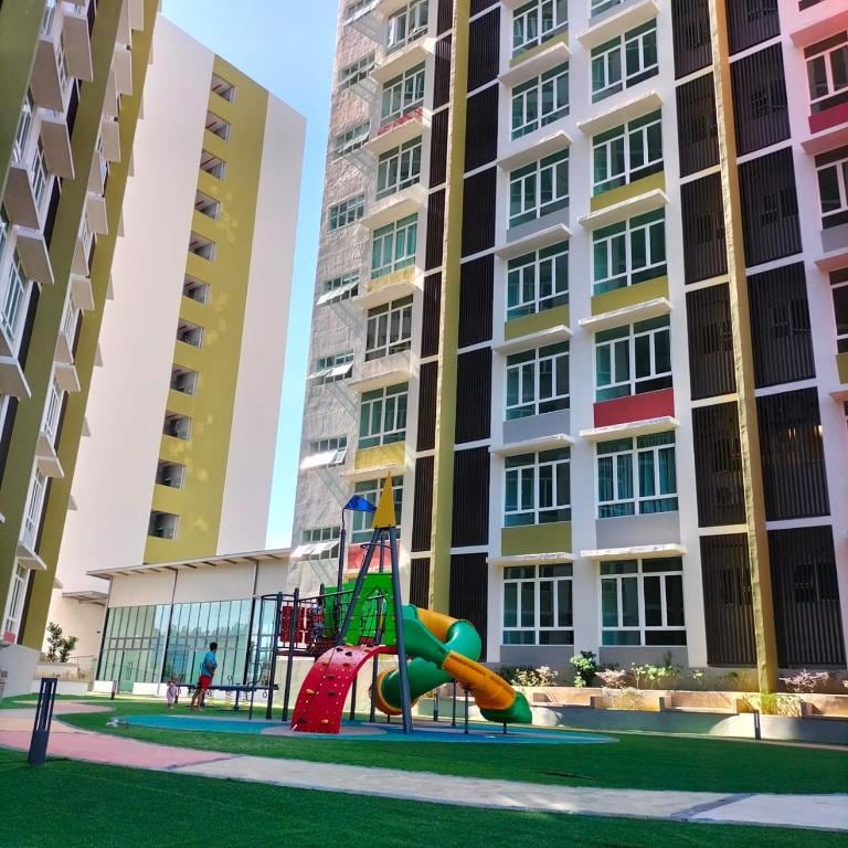 A colorful playground, featuring slides and climbing structures, is located in a grassy courtyard surrounded by modern apartment buildings. Large windows on the structures reflect natural light, creating a vibrant outdoor play area for families.