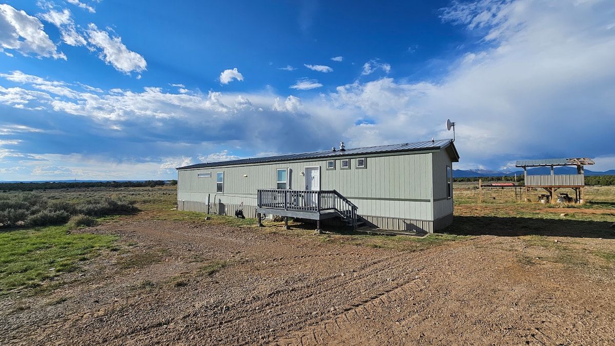 A single-story structure is set amidst an expansive outdoor area, with a wrap-around porch leading to the entrance. The building features a muted green exterior under a wide blue sky, with scattered clouds, and is surrounded by open fields and distant mountains.