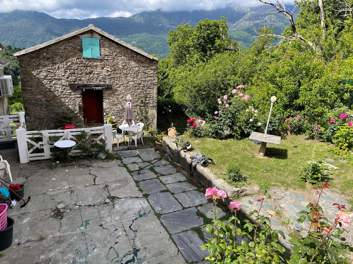 The exterior of a restored stone house is depicted, featuring a paved courtyard with a mix of stone and grass surfaces. Green foliage and blooming roses surround the area. Mountain views are visible in the background, creating a natural backdrop for relaxation.