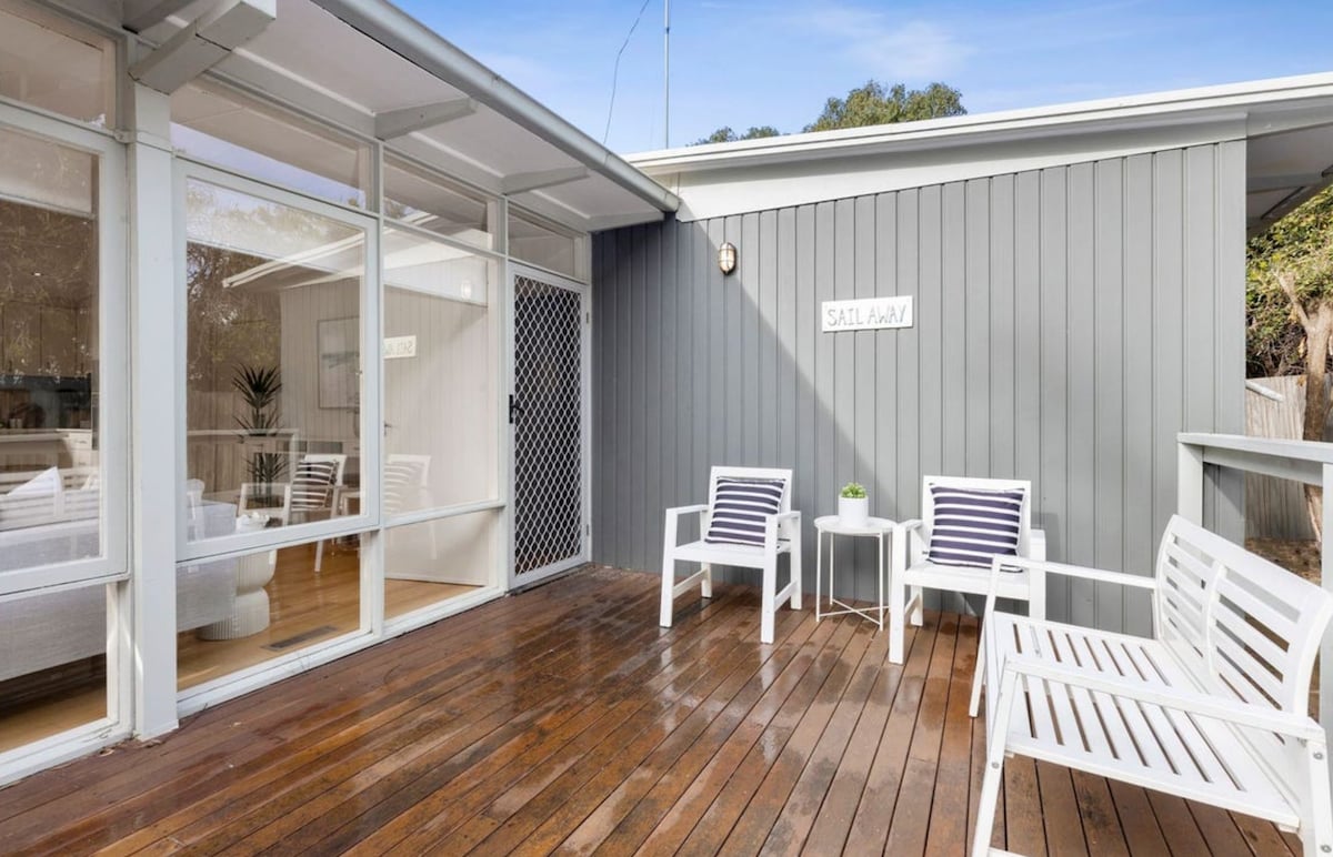 A spacious wooden deck features white outdoor seating with striped cushions, creating a comfortable space for relaxation. Light and shadow dance across the surface, while glass doors open to an interior, enhancing the connection to nature.