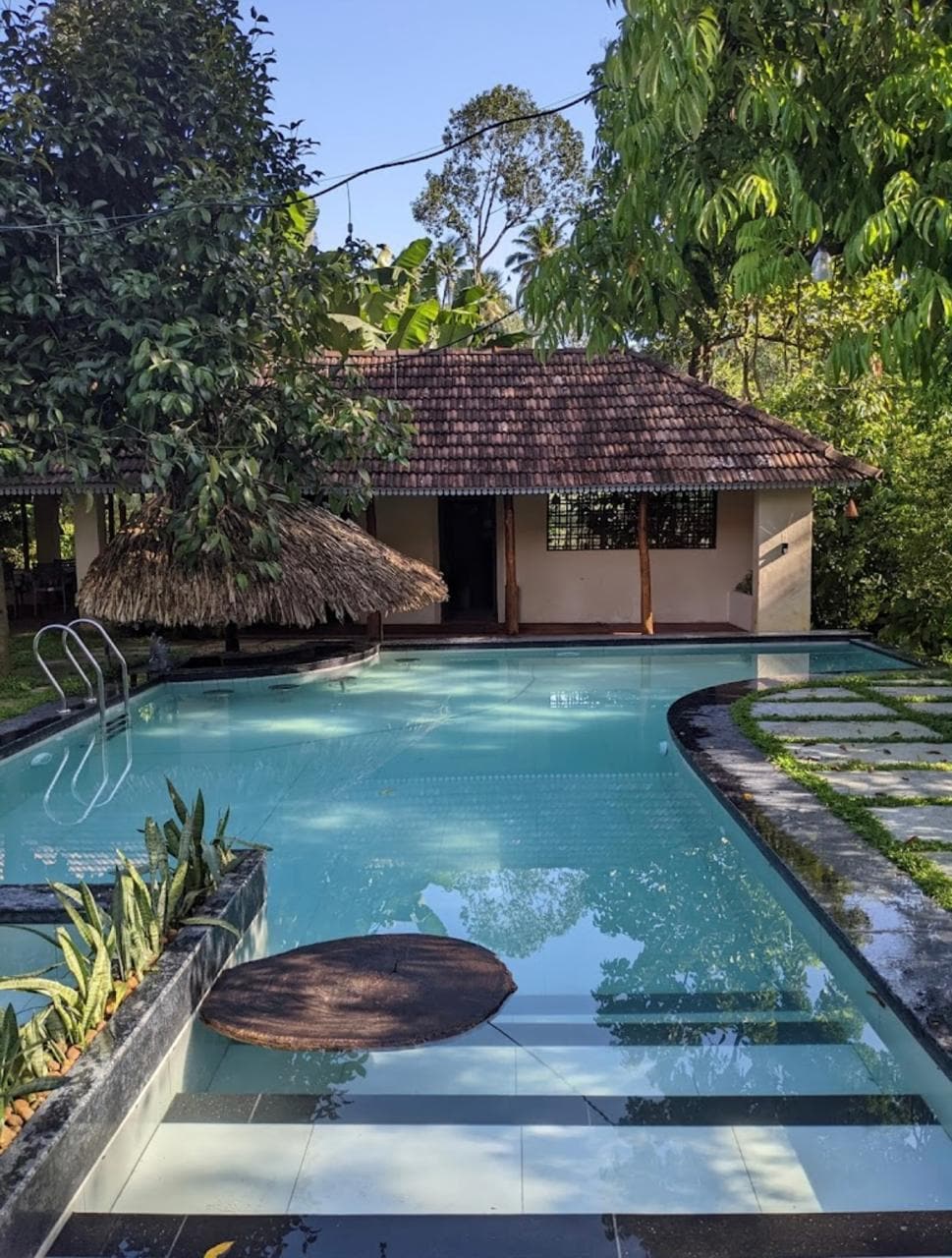 A serene swimming pool is framed by lush greenery and features a sleek stone pathway leading to it. The thatched-roof structure is visible in the background, providing shade and a rustic charm to the setting.