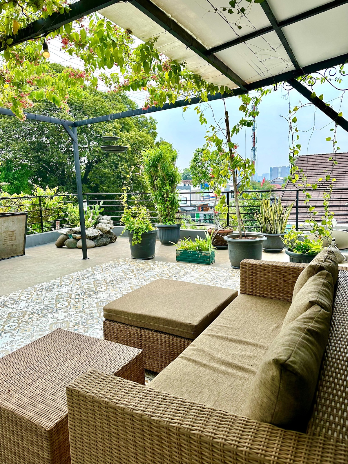 A spacious rooftop area is adorned with potted plants and greenery, providing a relaxing setting. Comfortable seating is arranged around a low table, while a canopy offers shade. The surrounding views of the city skyline are visible in the distance.