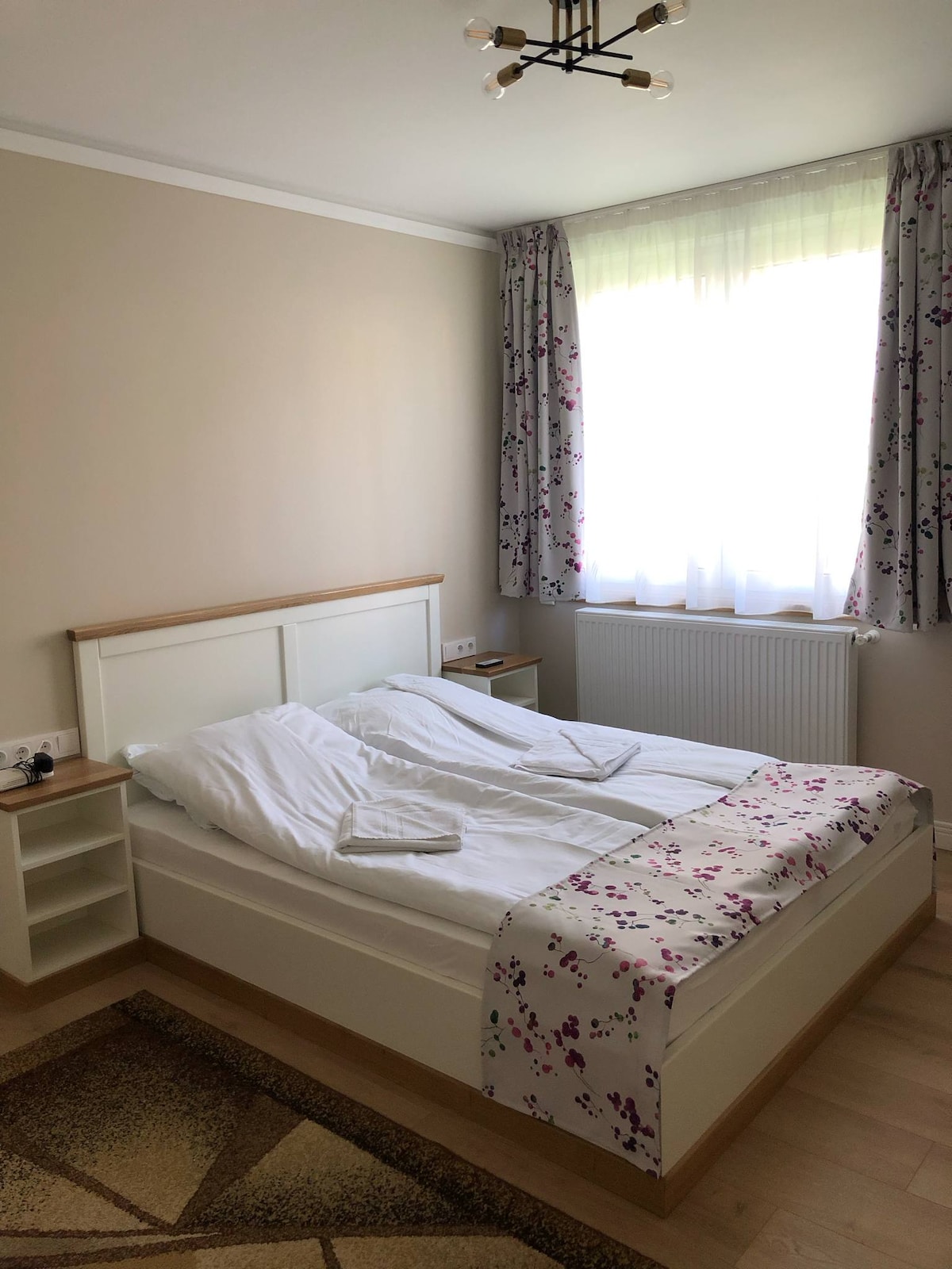 A bright bedroom features a spacious bed dressed in white linens and a floral-accented blanket. A large window allows natural light to fill the room, complemented by light curtains with floral patterns. A bedside table and radiator are also visible against a neutral wall.