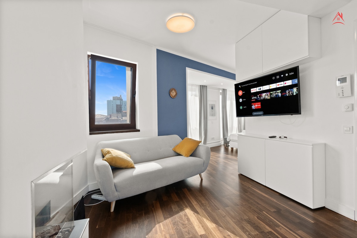 A contemporary living area features a light-colored sofa with a yellow cushion, positioned next to a sleek, white media unit. A large window offers a view of the cityscape, while neutral-colored walls complement the wooden flooring.