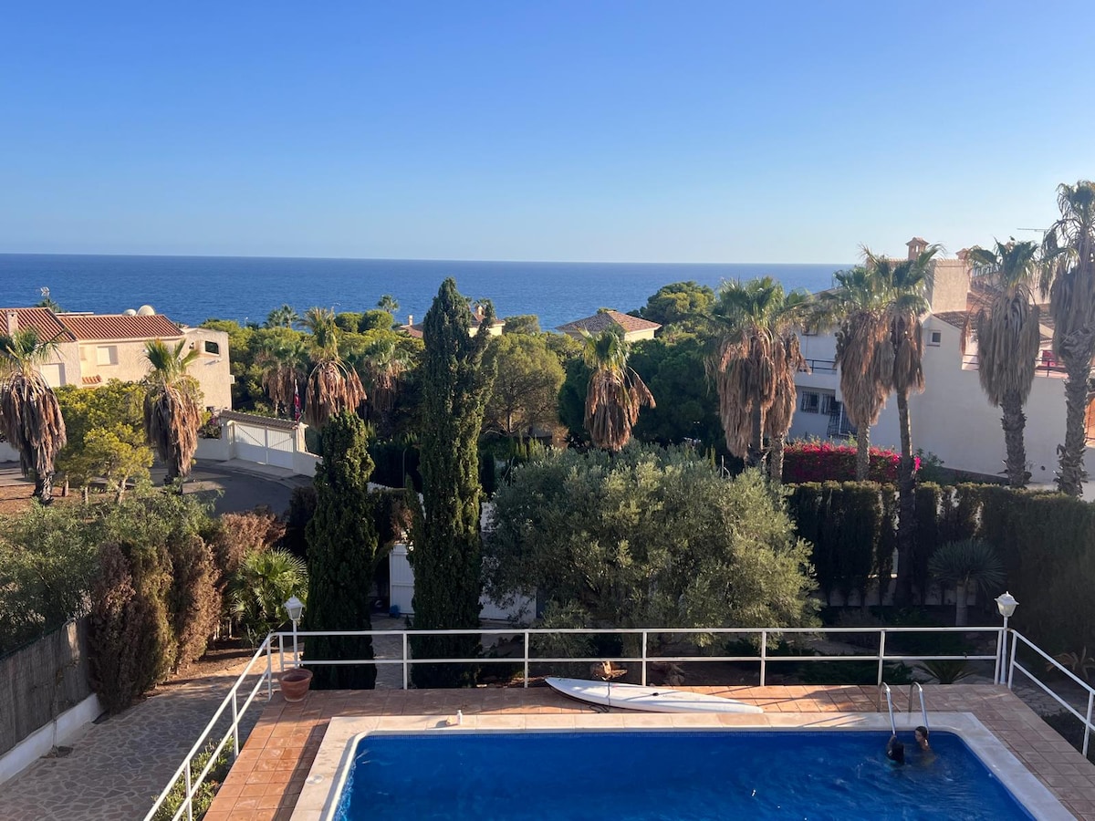 A tranquil scene features a swimming pool surrounded by lush greenery, with palm trees and shrubs visible in the background. The sparkling blue sea is seen beyond the property, illuminated by natural light under a clear sky. A serene coastal atmosphere is evoked.