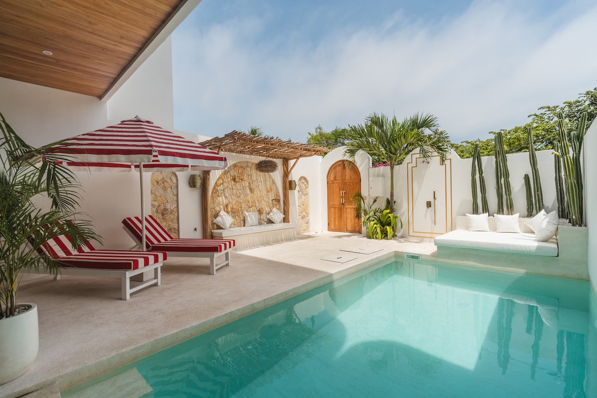 A serene outdoor space features a private pool surrounded by lush greenery. Two striped lounge chairs are positioned near the water, while a sunken couch with neutral cushions is tucked against the wall, offering a comfortable area for relaxation.