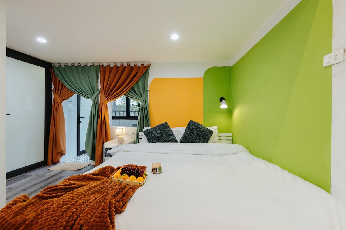 A bright bedroom presents a spacious bed adorned with a soft blanket, complemented by colorful wall accents in green and orange. Natural light enters through a window, with curtains in muted tones framing the view. Decorative pillows add a touch of comfort to the inviting space.
