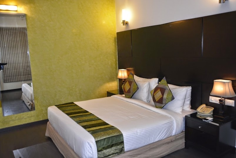 A modern bedroom features a spacious bed with white linens and decorative pillows. Soft lighting from bedside lamps illuminates the room. A vibrant green accent wall complements the dark headboard, while a mirrored wall reflects the space, enhancing its brightness and openness.