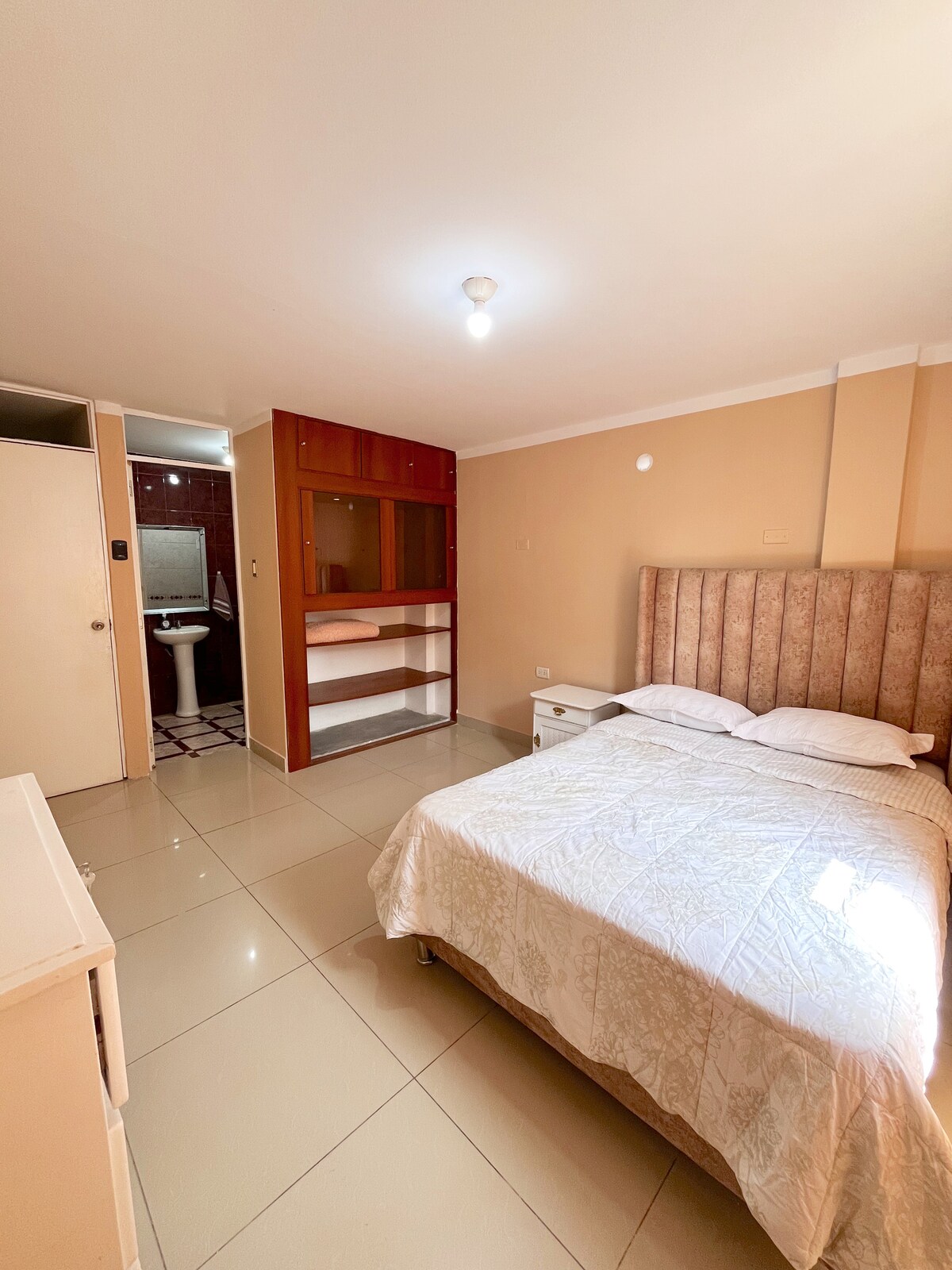 A comfortable bedroom is highlighted by a spacious bed draped with a light-colored coverlet. Storage space is provided by a wooden shelf and a dresser. A private bathroom is visible in the background, featuring essential fixtures and a tiled wall.