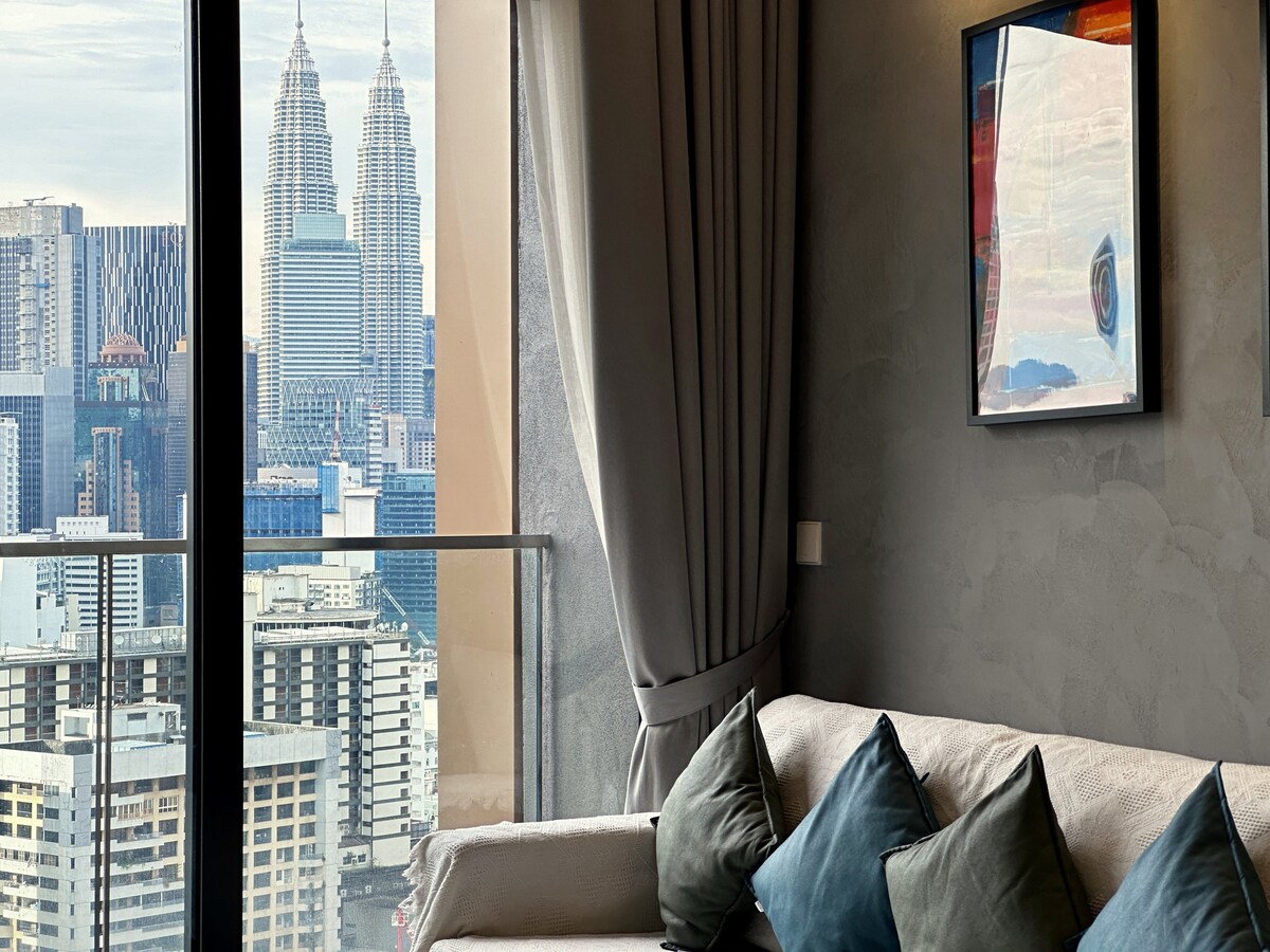 A spacious living area showcases a comfortable sofa with decorative cushions, positioned near large glass doors. The view features the iconic Petronas Twin Towers rising amidst the Kuala Lumpur skyline, framed by soft curtains and a piece of modern art on the wall.