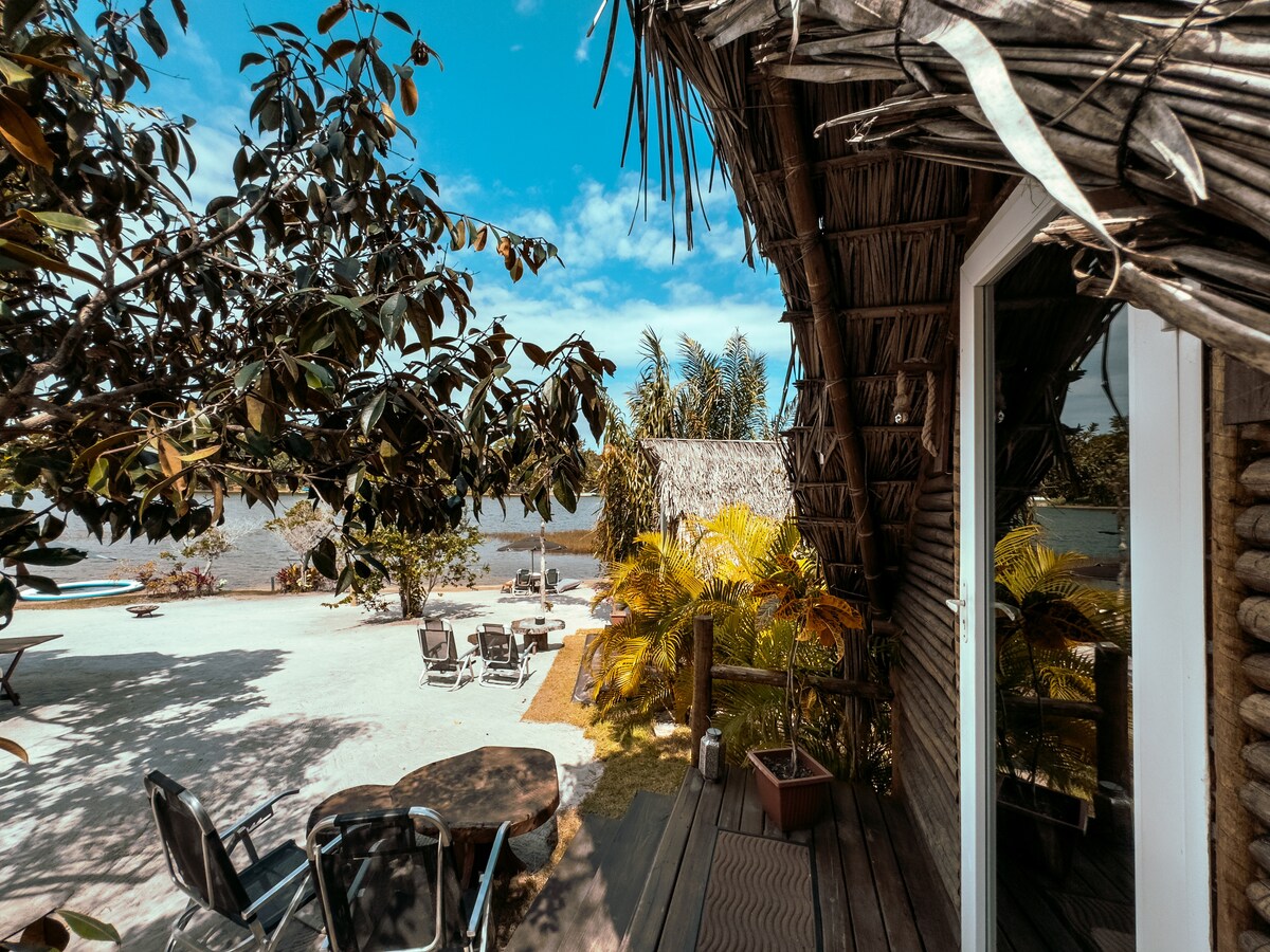 A view from the cabana showcases lush greenery and inviting outdoor furniture. The scene features a sandy area with scattered lounge chairs and a table, complemented by tropical plants. The tranquil lagoon is visible in the background under a bright blue sky.