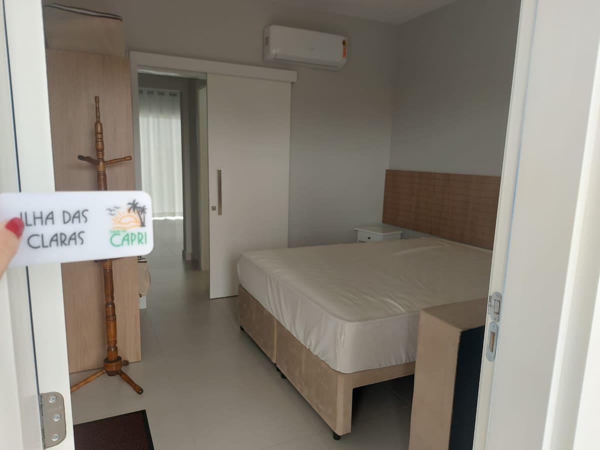 A spacious bedroom features a queen bed, light-colored walls, and a minimalist design. A nightstand is positioned beside the bed, and natural light enters through a window dressed with sheer curtains. An air-conditioning unit is mounted on the wall for climate control.