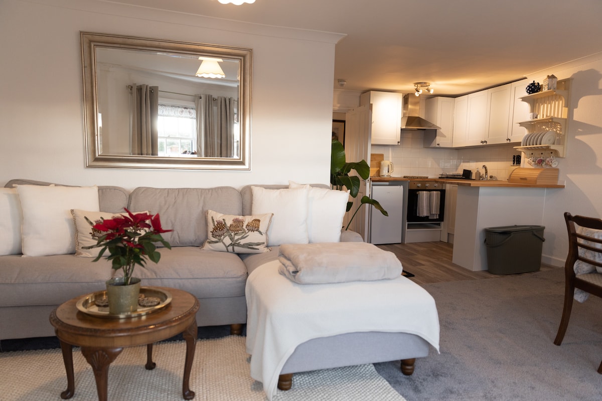A comfortable sitting area features a light-colored sofa adorned with decorative pillows. A small round table holds a potted plant, while an ottoman is positioned nearby. The open-plan kitchen is visible in the background, showcasing modern appliances and light cabinetry.