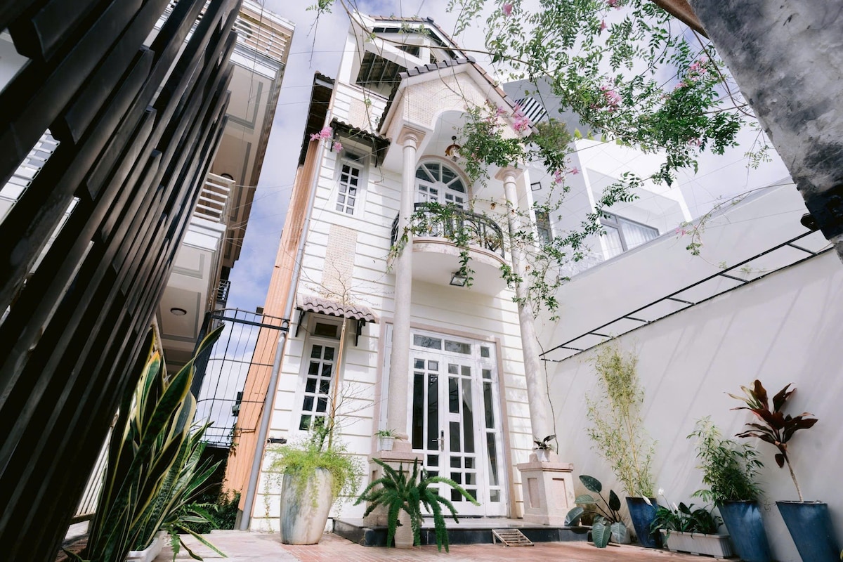 The exterior of a two-story building is depicted, featuring tall windows and a prominent balcony with decorative railings. Lush greenery surrounds the entrance, complemented by potted plants positioned on the ground. Sunlight illuminates the façade, which showcases intricate architectural details.