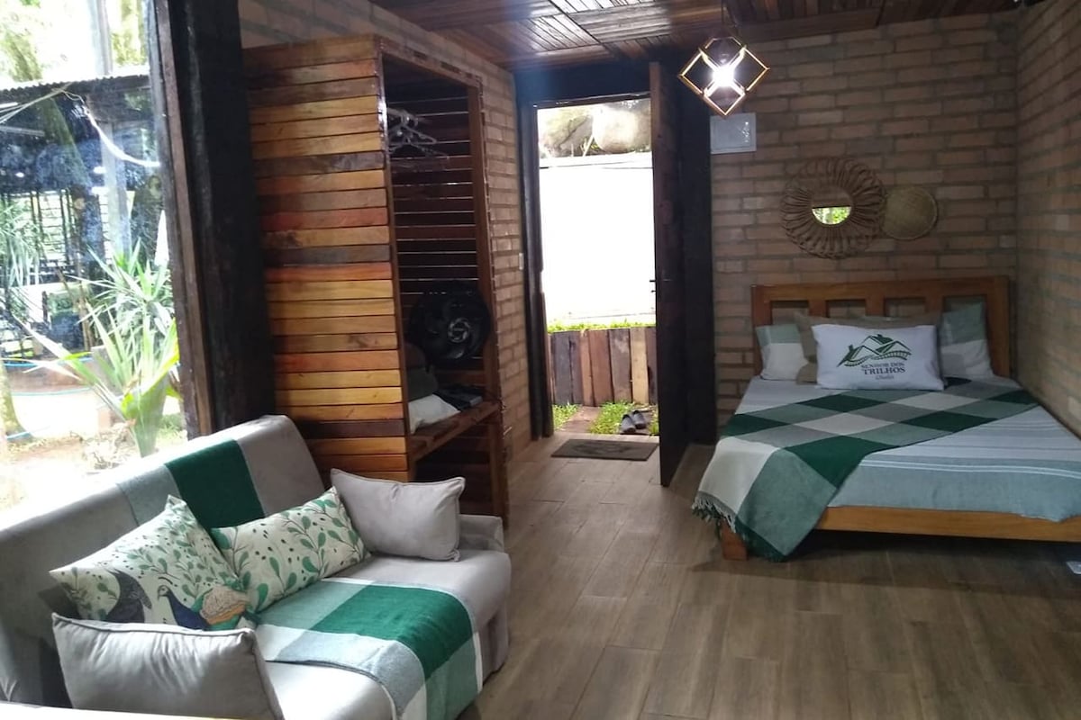 A cozy interior space is presented, featuring a wooden bed topped with a green and white blanket. Nearby, a sofa with decorative pillows is positioned, enhancing the inviting layout. Natural light filters in through the door leading to the outdoor area, complementing the rustic brick walls.