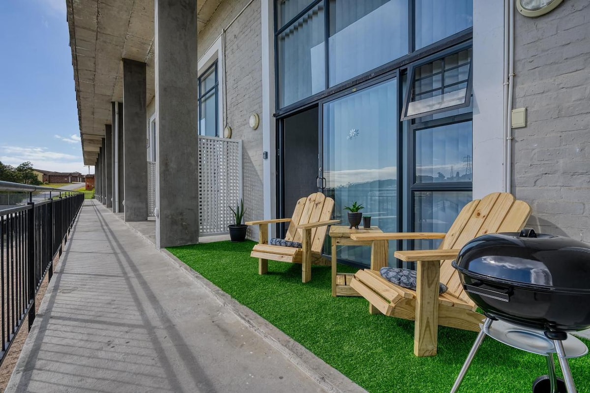 A balcony area features two wooden chairs on artificial grass, offering a comfortable setting. A small BBQ grill is positioned nearby, creating an inviting space. Large glass doors provide access to natural light, while planters add greenery to the environment.