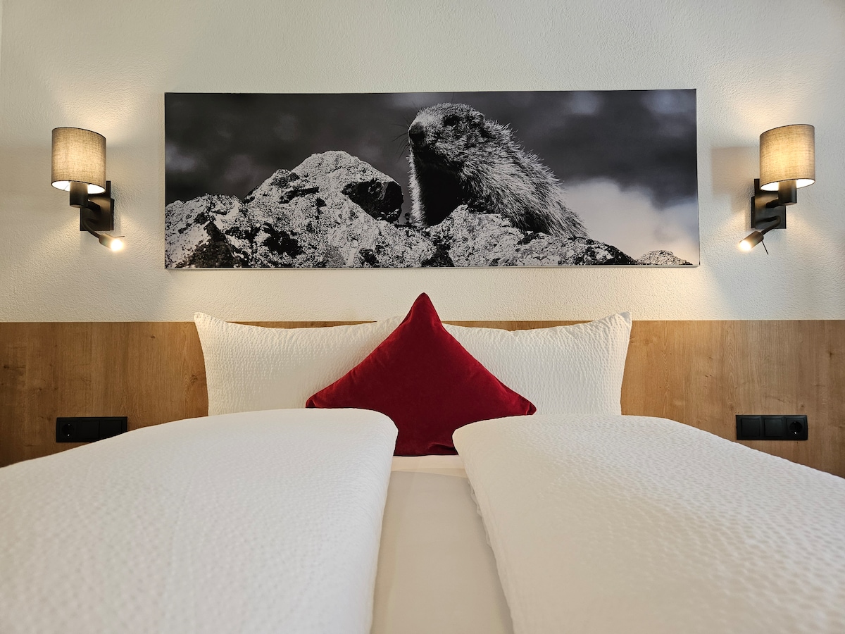 The sleeping area features a spacious bed adorned with soft linens. A prominent red accent pillow adds contrast. On the wall above, a large black-and-white photograph of a mountain animal is displayed. Wall sconces on either side provide gentle illumination.