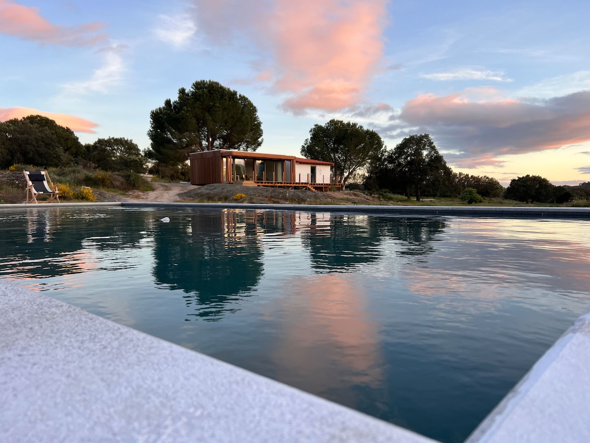 A serene outdoor scene captures a tranquil swimming pool reflecting the sky's pastel hues. In the background, a modern house is framed by lush greenery, creating a harmonious connection with nature.