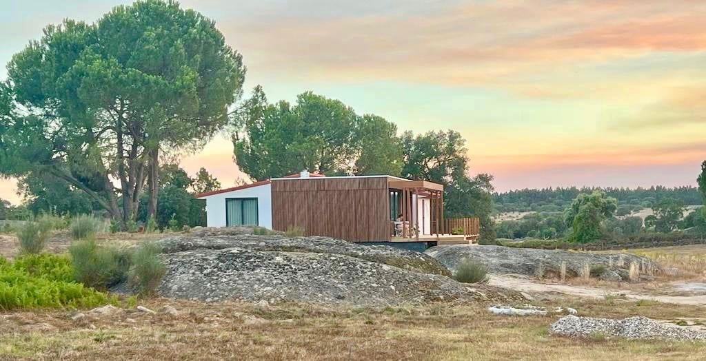 A modestly designed cottage sits nestled among trees, with a warm sunset casting soft hues across the sky. The structure features large windows, blending nature with comfort, while surrounding grassy areas enhance the tranquil rural setting.