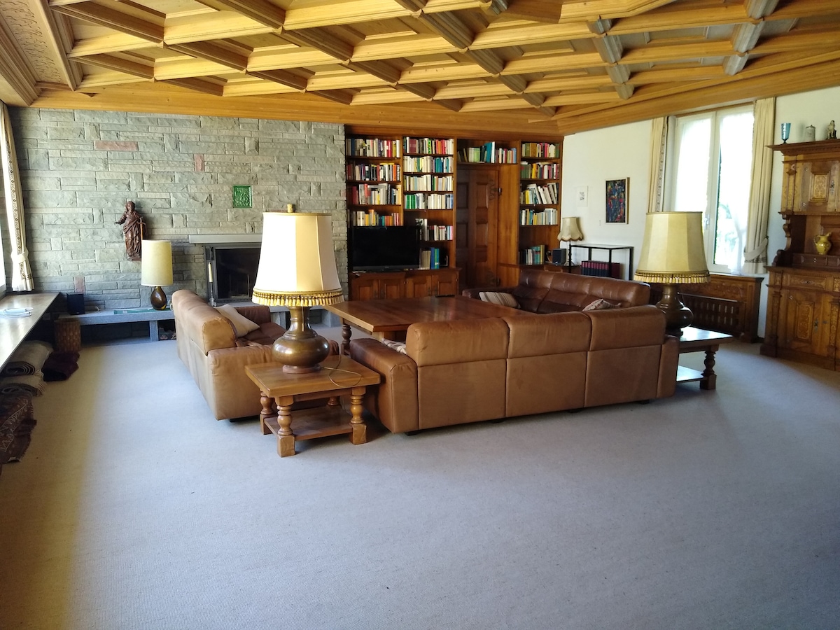 A spacious living area features a large leather sectional sofa centered around a coffee table, complemented by two lamps. The ceiling showcases wooden beams, and a stone wall adds texture. A bookshelf filled with books lines the back wall, providing an inviting backdrop.