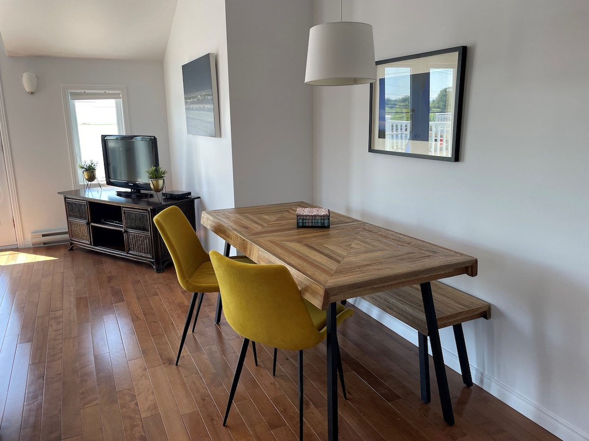 A wooden dining table with a chevron pattern is accompanied by two yellow chairs. The space features a television and a low cabinet against the wall, with framed artwork enhancing the modern décor. Natural light brightens the area through a nearby window.