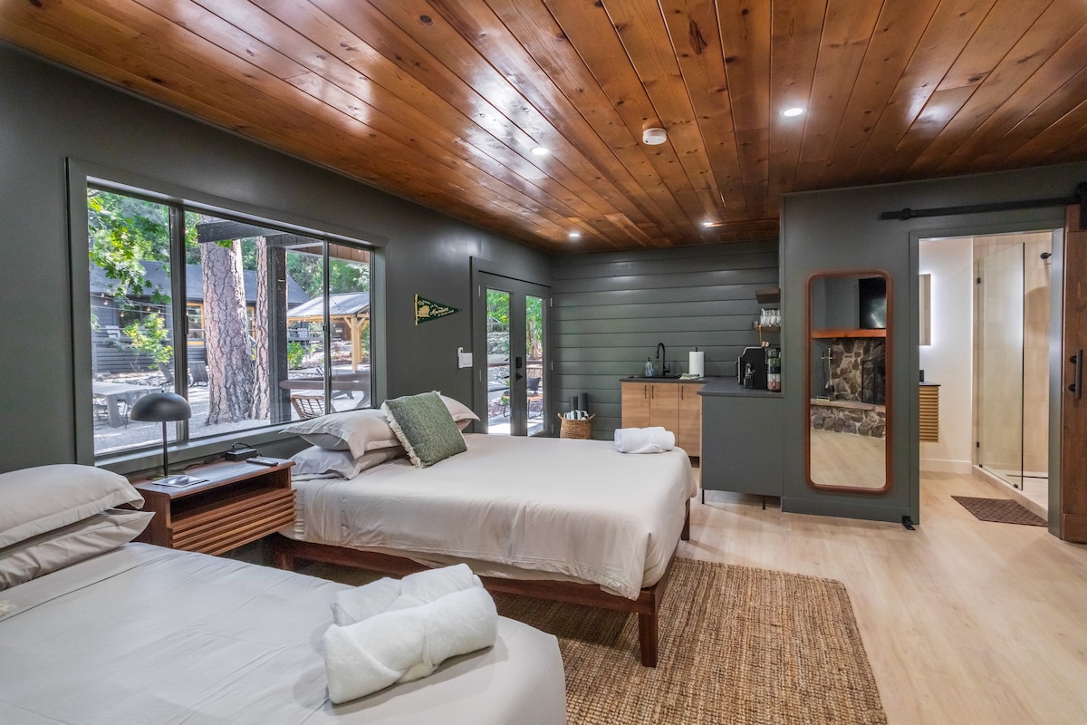 The spacious room features two queen beds, complemented by natural wood accents and a large window offering views of the outdoor courtyard. A kitchenette is visible with essential appliances, while a large tiled bathroom is accessed through a barn door.