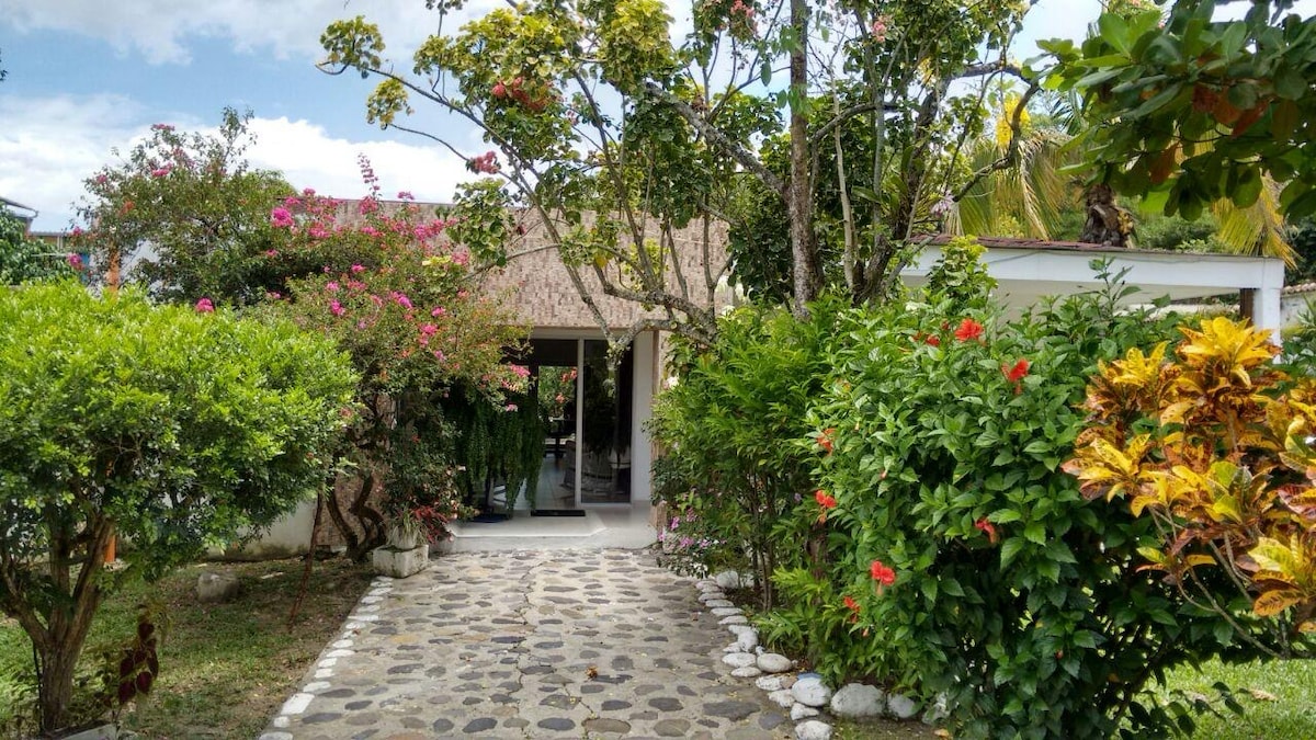 A stone pathway leads through vibrant greenery and flowering plants to the entrance of a house. The building features a thatched roof and large glass doors, blending harmoniously with the surrounding tropical landscape under a partly cloudy sky.