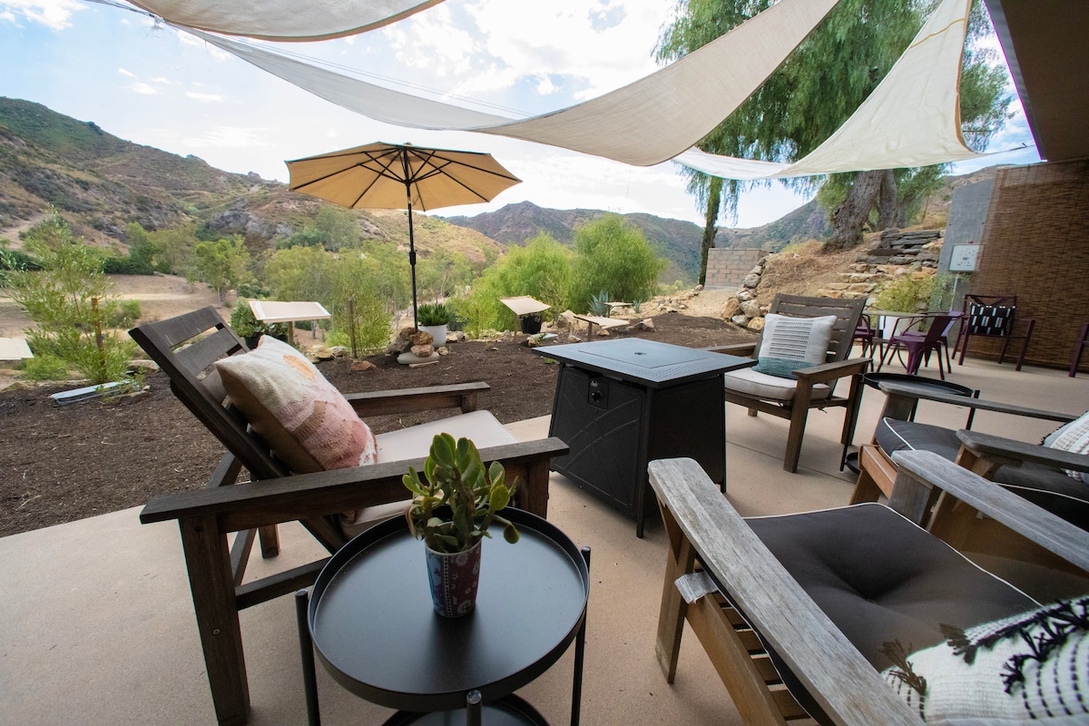 A private patio area features comfortable lounge chairs and a small table, inviting relaxation. An umbrella offers shade from the sun, while the landscape reveals scenic mountain views and greenery in the background. A fire pit is centrally located, enhancing outdoor enjoyment.