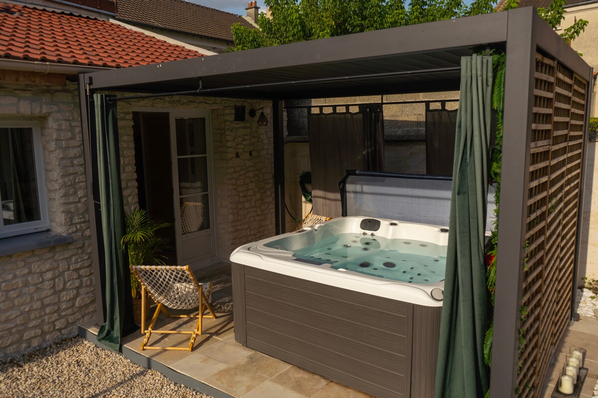 A private outdoor spa area is enclosed by a stylish structure featuring wooden slats. A hot tub is positioned within the space, complemented by two comfortable seating options under the shade. Lush greenery surrounds the area, enhancing the serene environment.