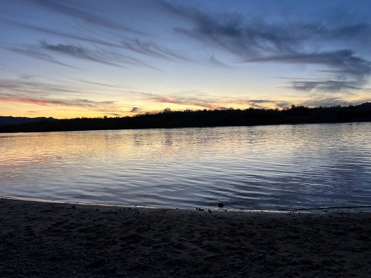 A serene sunset over a calm river is captured, with vibrant hues of orange and purple reflected on the water's surface. The sandy shoreline gently leads to the water's edge, and silhouettes of distant land are visible in the background.