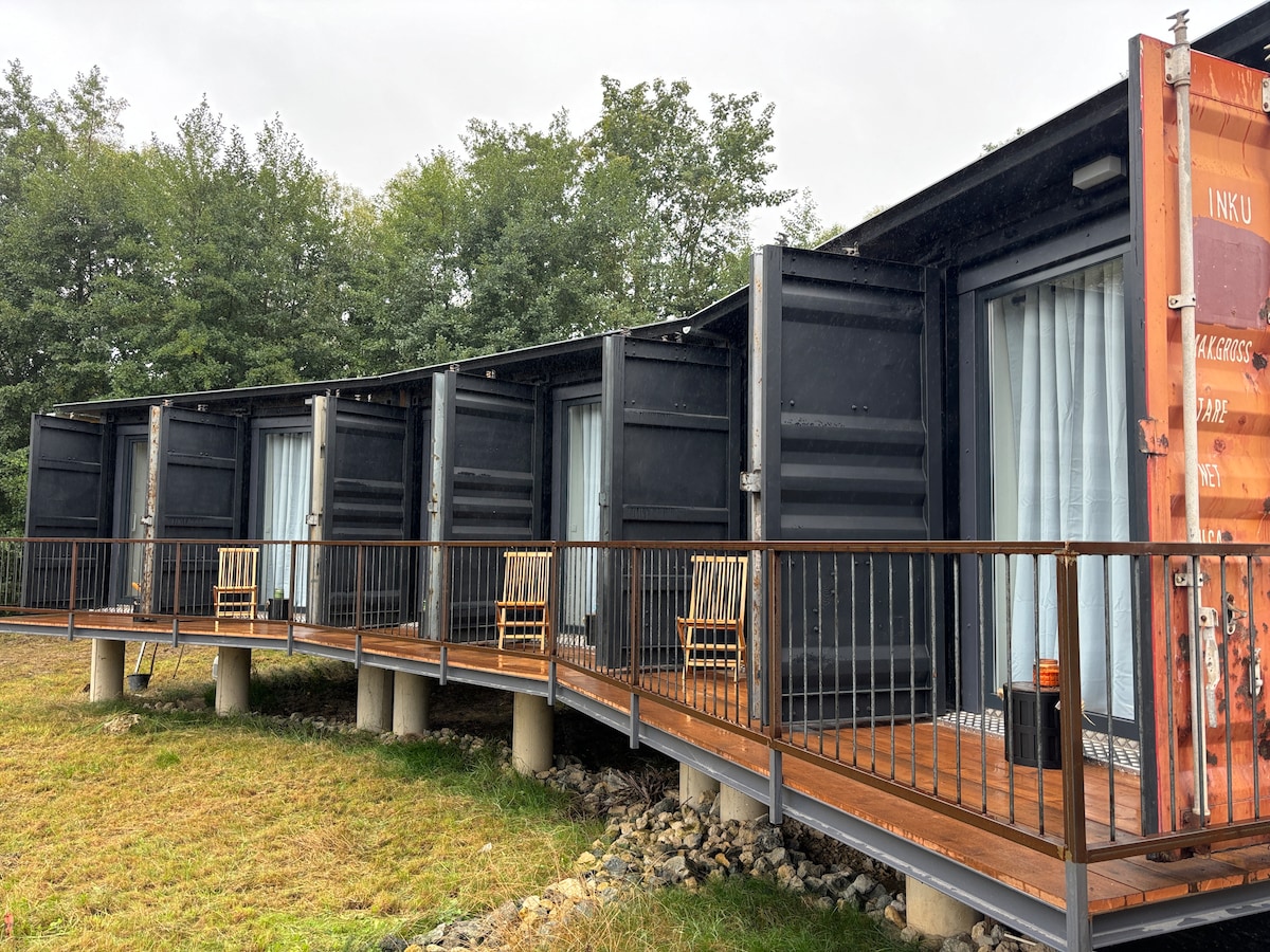 Multiple stylishly converted shipping containers are arranged in a row, each with sliding glass doors. A wooden deck with railing connects the containers, featuring seating areas with chairs positioned to enjoy the surrounding green landscape.