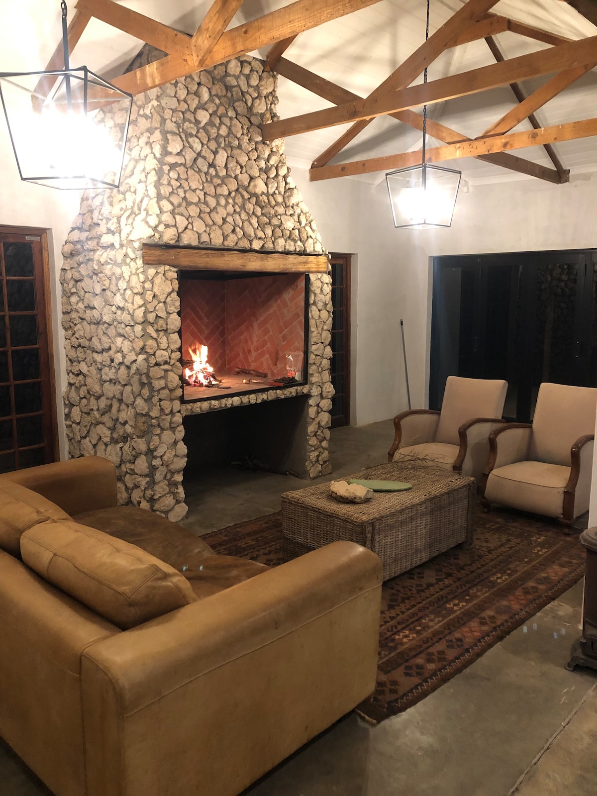 A cozy living area is highlighted by a stone fireplace with a warm fire. Comfortable seating is provided by two armchairs and a leather sofa, while a coffee table sits in the center. The room is softly illuminated by pendant lights and has a relaxing color palette.