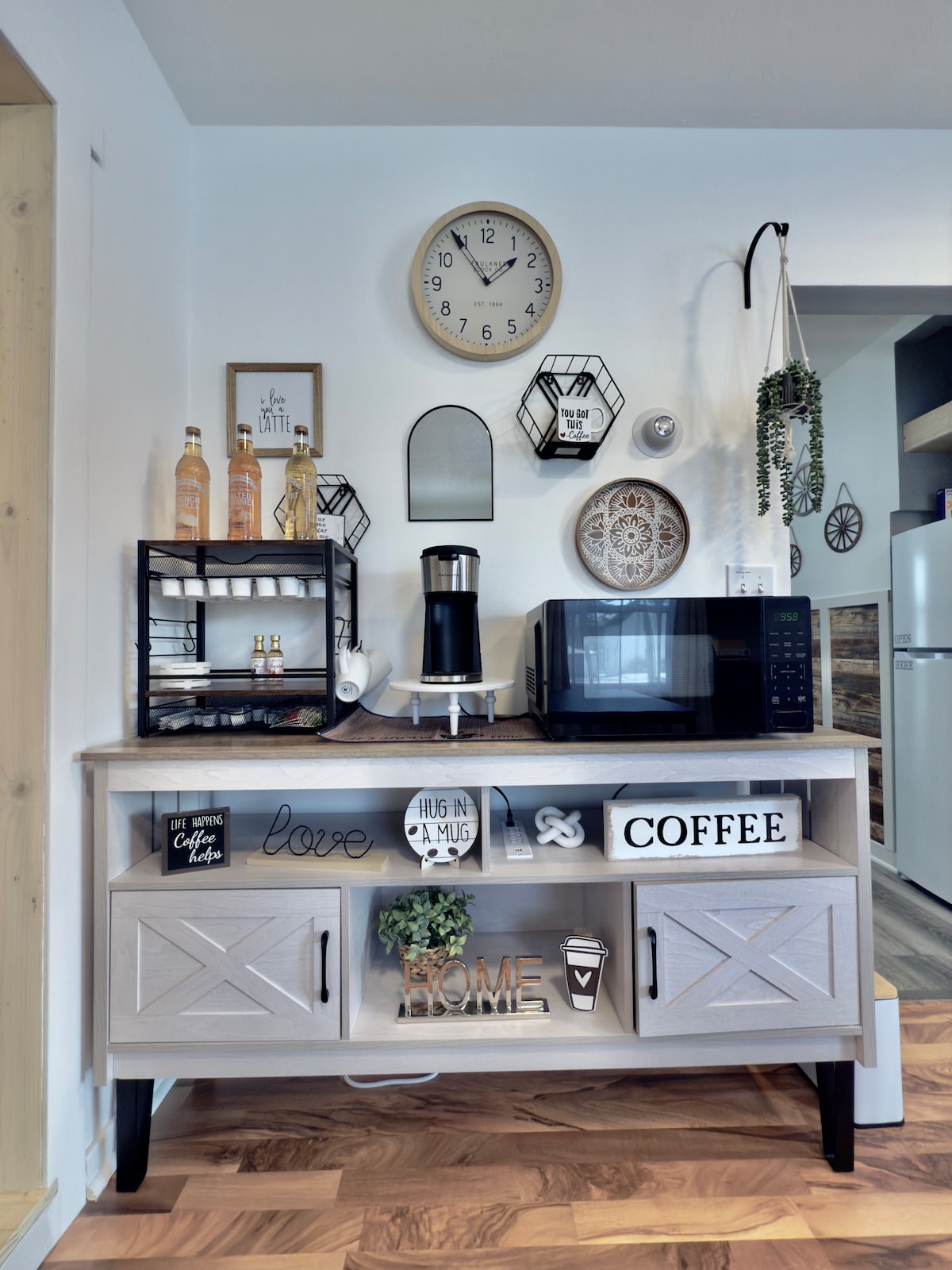A modern kitchen area is showcased, featuring a wooden cabinet with decorative elements. A coffee maker, microwave, and various bottles are neatly arranged on the countertop. Wall decor includes a clock, mirrors, and framed art, enhancing the functional space.